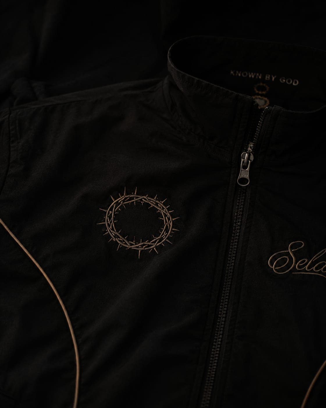 Stillness Track Jacket in black — tonal crown of thorns embroidery, KNOWN BY GOD collar, leather-brown piping